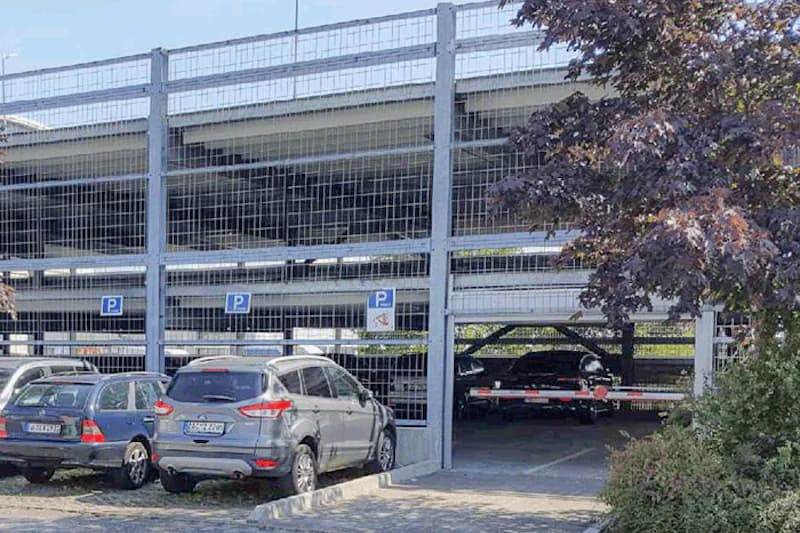Mehrstöckiges vergittertes Parkhaus mit geparkten Autos und sichtbarer Einfahrtsschranke, Baum rechts im Vordergrund.