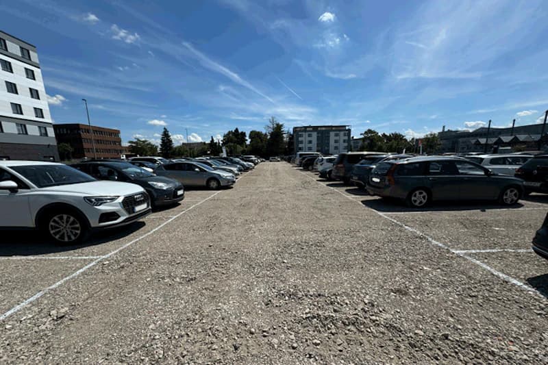 Schotterparkplatz mit geparkten Autos in Reihen und offener Fahrspur in der Bildmitte.