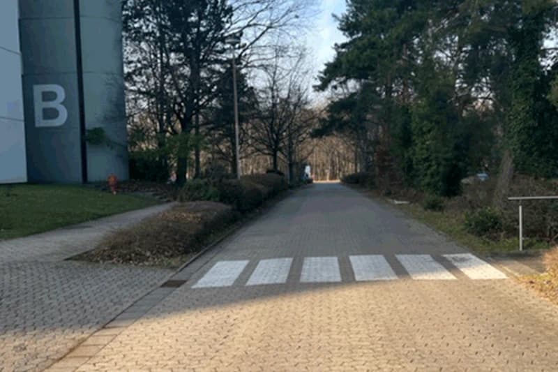 Gepflasterter Weg mit Zebrastreifen, Gebäude mit großem B links, Bäume und Sträucher beidseits.
