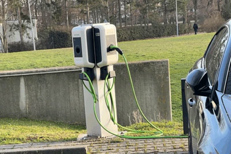 Ladesäule mit grünen Ladekabeln neben geparktem Auto auf einem Parkplatz.
