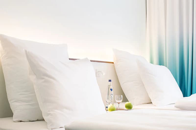 Zwei weiße Kissen auf einem Bett mit Wasserflasche, Glas und Äpfeln am beleuchteten Kopfteil und türkisfarbener Gardine.