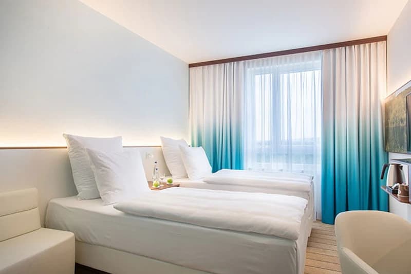 Zweibettzimmer mit zwei Einzelbetten, weißen Bettwaren, LED-Lichtleiste, türkisverlaufenen Vorhängen und Fenster.