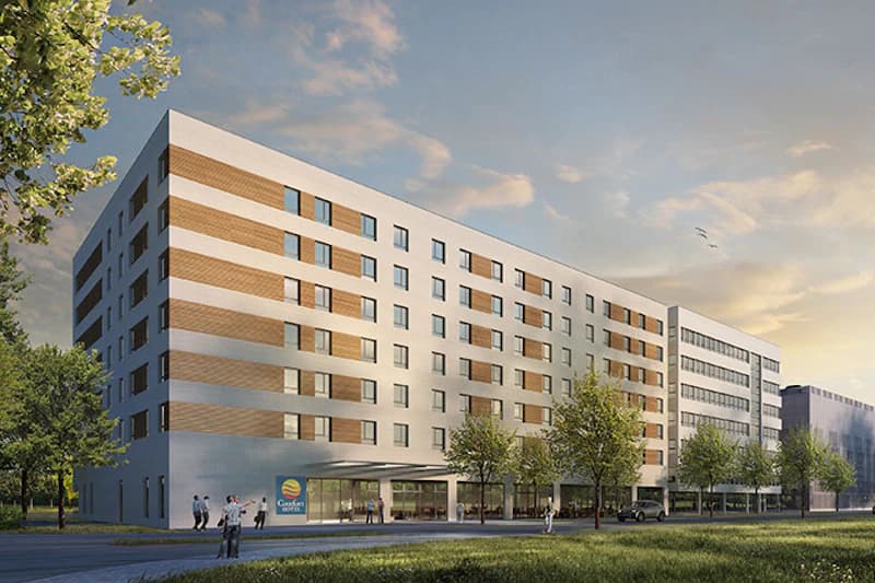 Mehrstöckiges Hotelgebäude mit gleichmäßigen Fenstern, Vordach am Eingang, Bäumen davor und Personen auf dem Gehweg.