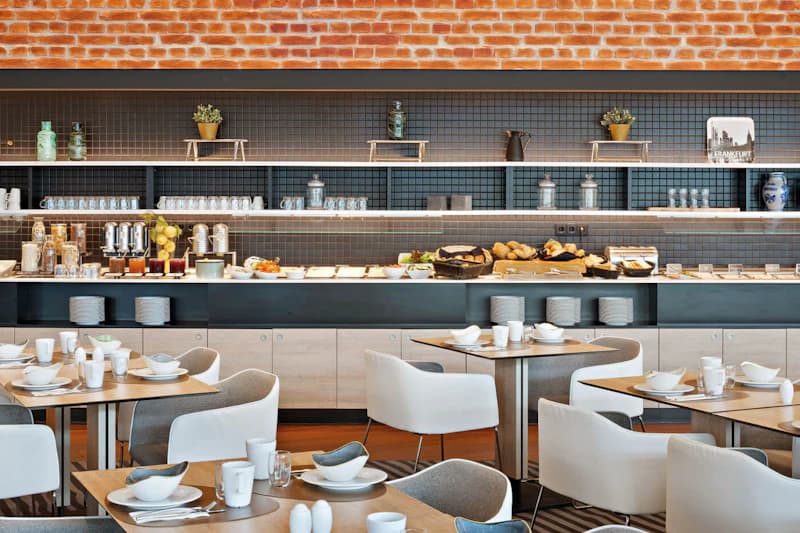 Frühstücksbuffet mit gedeckten Tischen, Sesseln und Buffetzeile mit Backwaren und Geschirr vor Ziegelwand.
