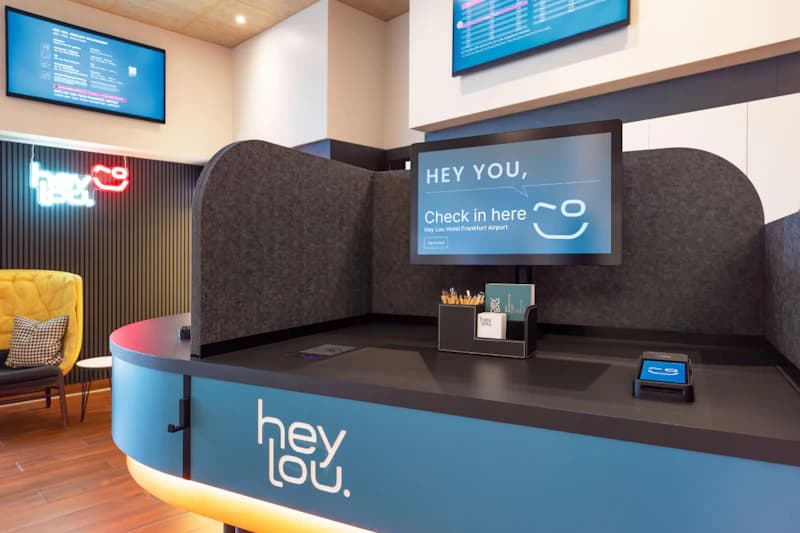 Empfangstheke des Hey Lou Hotels mit blauem Logo, Monitor, grauen Trennwänden und gelbem Sessel im Wartebereich.