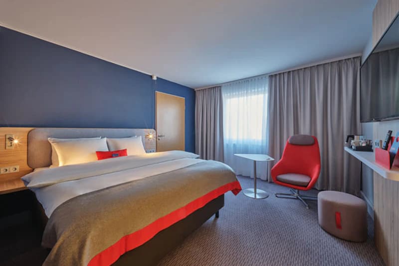 Doppelzimmer mit großem Bett vor blauer Wand, Fenster mit Vorhang, rotem Sessel, Pouf, Wandfernseher und kleinem Tisch.