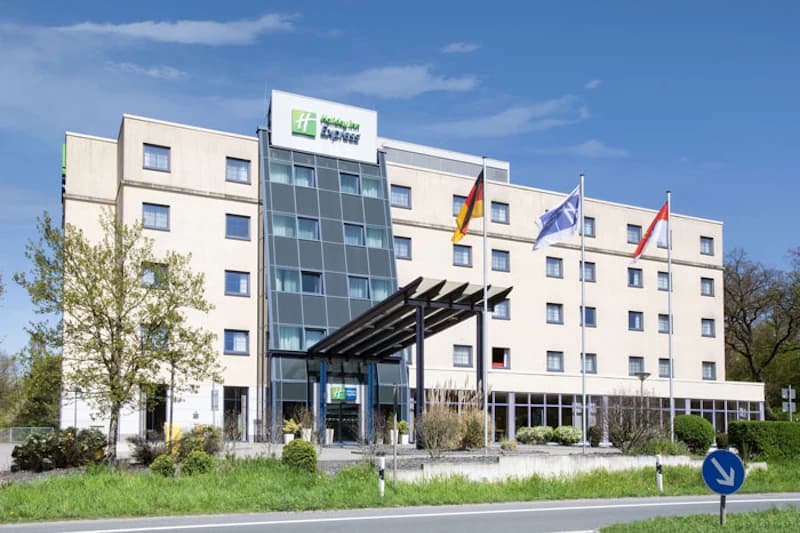 Mehrstöckiges Hotelgebäude mit gläsernem Mittelteil, Eingangsüberdachung, drei wehenden Fahnen und Verkehrszeichen.