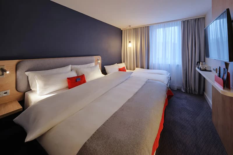 Hotelzimmer mit zwei Betten, dunkler Akzentwand, Wandfernseher, Vorhang und Nachttisch mit Leselampen.