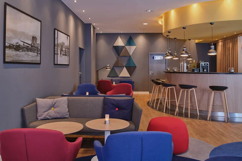 Hotel-Lounge mit Sofas, bunten Sesseln, Couchtischen und Bartresen mit Hockern und Hängeleuchten.