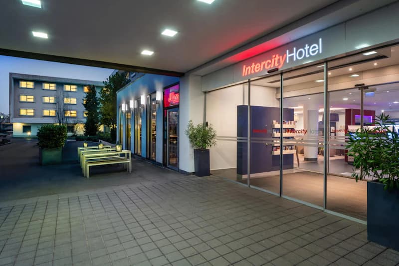 Eingangsbereich des IntercityHotel mit beleuchtetem Schriftzug, Glastüren und Außensitzbänken bei Dämmerung.