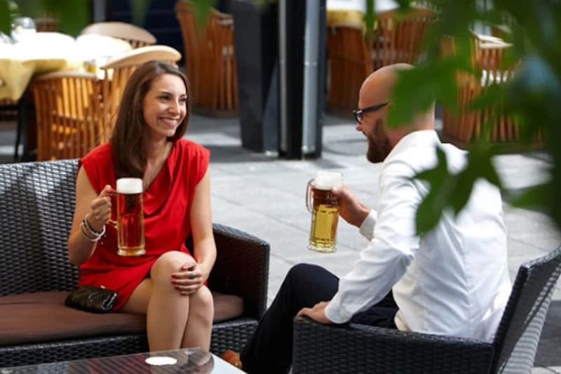 Frau im roten Kleid und Mann im weißen Hemd sitzen auf Lounge-Sofas und stoßen mit Bierkrügen an.
