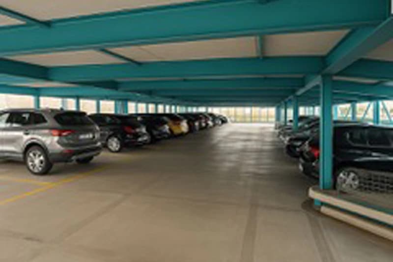 Überdachtes Parkdeck mit geparkten Autos, türkisfarbene Stahlträger und breite Fahrspuren.