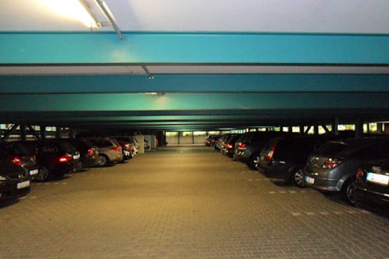 Überdachtes Parkdeck mit niedriger Decke, zwei Reihen geparkter Autos und zentraler Fahrspur, künstlich beleuchtet.