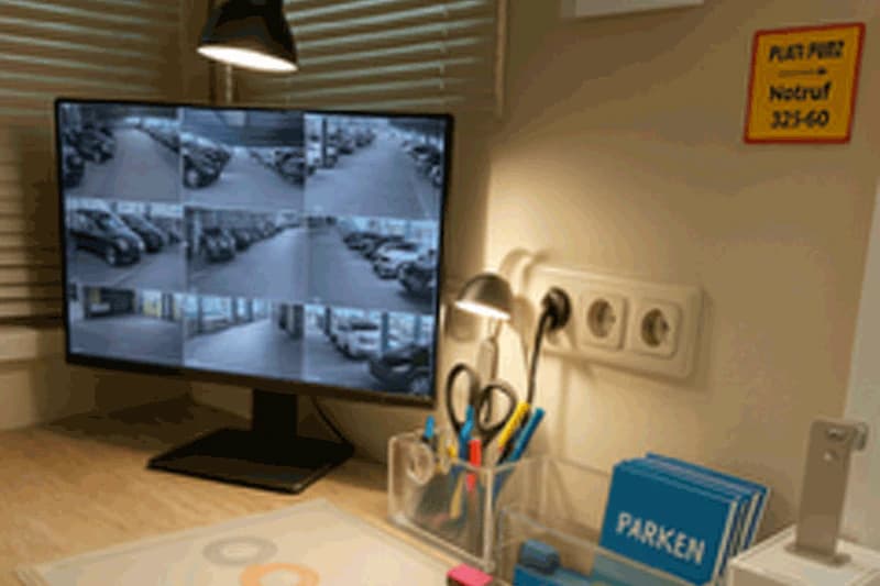 Überwachungsmonitor mit neun Kamerabildern eines Parkdecks, Schreibtischlampe und Stifthalter mit Parken-Heftchen.