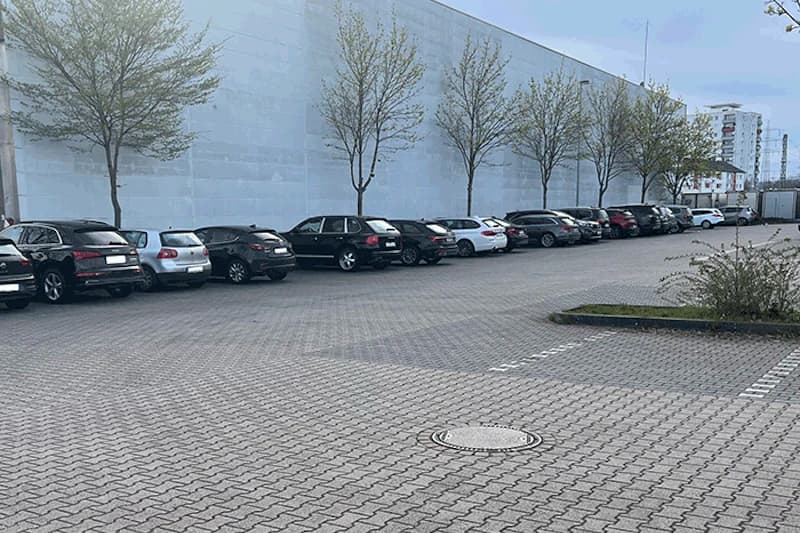 Reihen geparkter Autos an einer hohen weißen Hallenwand, gepflasterter Parkplatz mit Bäumen und Kanaldeckel.