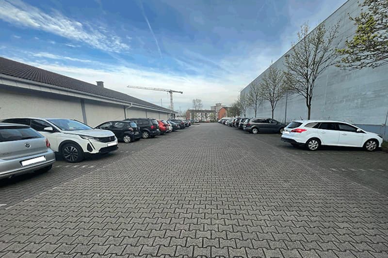 Gepflasterter Außenparkplatz mit Autos links und rechts, Gebäudewänden, Bäumen und einem Baukran am Horizont.
