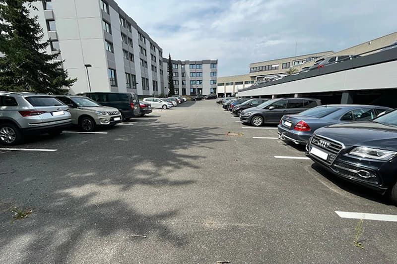 Offener Parkplatz zwischen mehrgeschossigen Gebäuden mit Reihen geparkter Autos links und rechts.