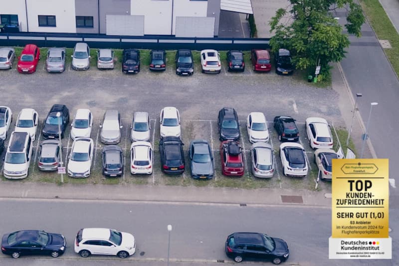 Luftaufnahme eines Freiluftparkplatzes mit mehreren Reihen geparkter Autos, Gebäude im Hintergrund und Straße vorne.