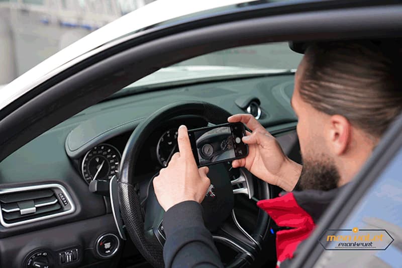 Mann sitzt am Lenkrad und fotografiert mit Smartphone das Armaturenbrett eines Autos.