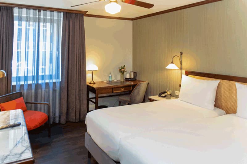 Zimmer mit zwei Einzelbetten, Nachttischlampe, Schreibtisch, Sessel, Fenster mit Vorhang und Deckenventilator.