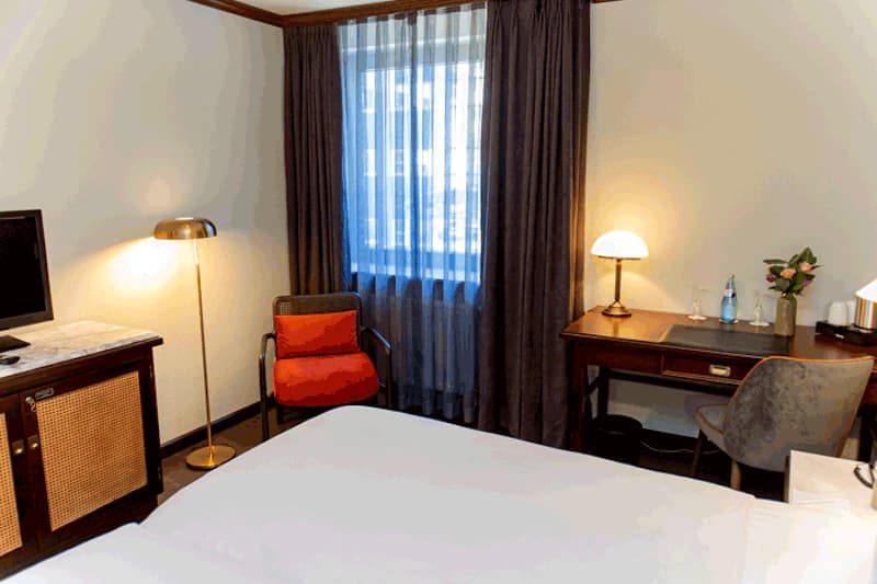 Zimmer mit Bett im Vordergrund, Fenster mit Vorhängen, roter Sessel, Schreibtisch mit Lampe, Fernseher und Blumen.