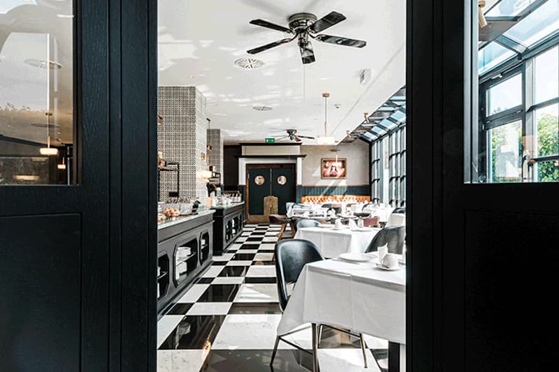 Hotelrestaurant mit schwarz-weiß kariertem Boden, gedeckten Tischen, Buffetzeile, großen Fenstern und Deckenventilatoren.