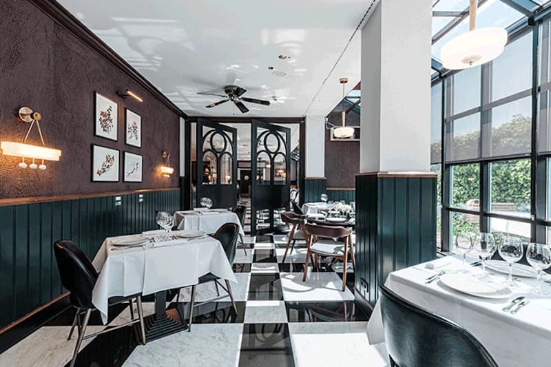 Restaurant mit schwarzweiß kariertem Boden, gedeckten Tischen, großen Fenstern und dunklen Holzverkleidungen.