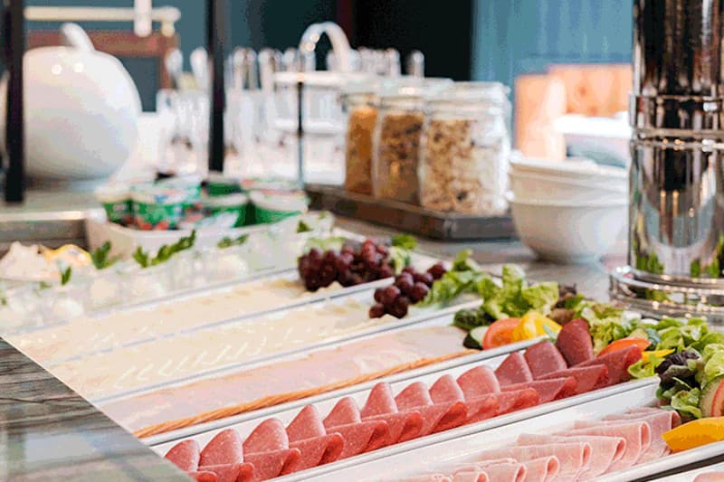Buffet mit aufgereihten Wurst- und Käsescheiben, Trauben, Salat und Gläsern mit Müsli beim Frühstück.