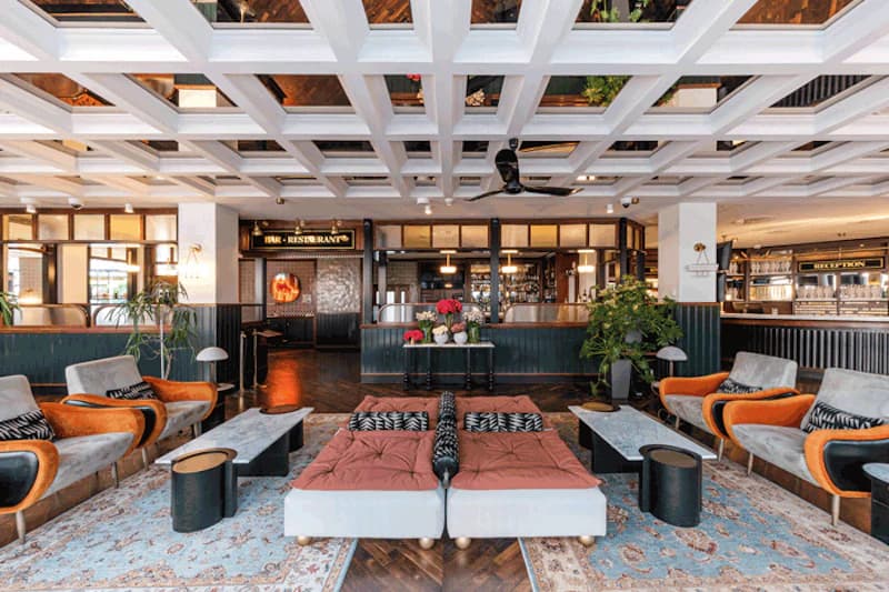 Hotellobby mit Sitzgruppen, niedrigen Tischen, Teppichen und sichtbarer Bar sowie Empfangsbereich im Hintergrund.