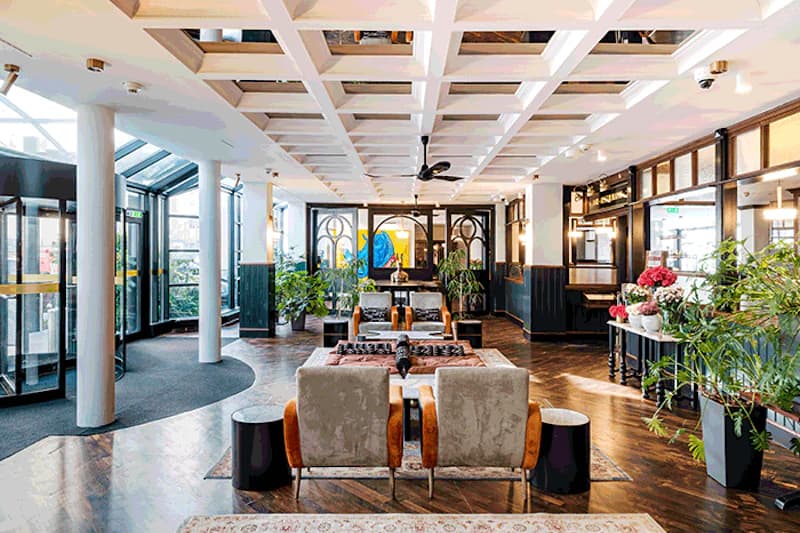 Hotellobby mit Sitzgruppen, Glasdrehtür links, Pflanzen, Holzfußboden und dekorativem Deckengitter.