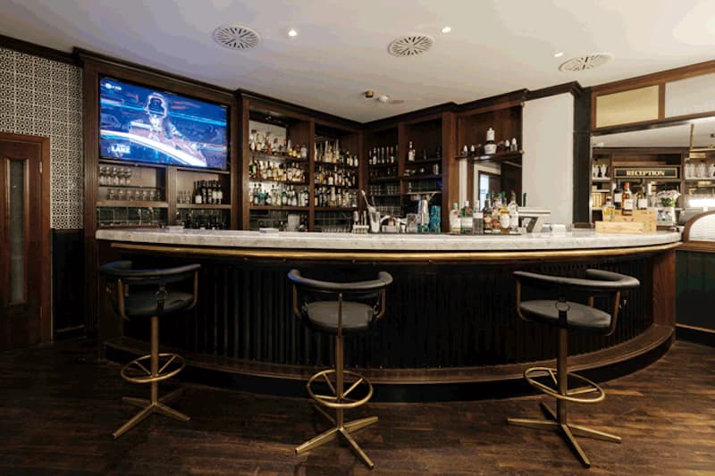 Halbrunde Hotelbar mit Marmorplatte, drei Barhockern und Regalen mit Flaschen sowie Fernseher an der linken Wand.