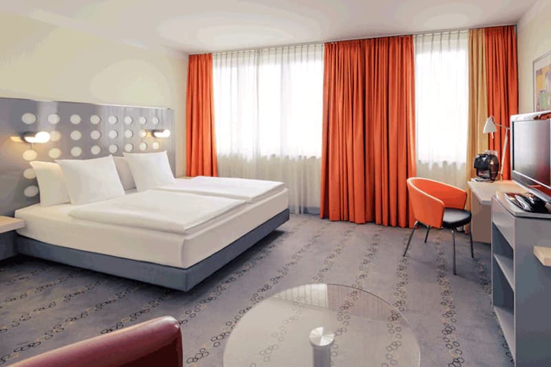 Helles Hotelzimmer mit Doppelbett, Kopfteil mit Punktmuster, orange Vorhängen, Schreibtisch, Fernseher und Kaffeemaschine.