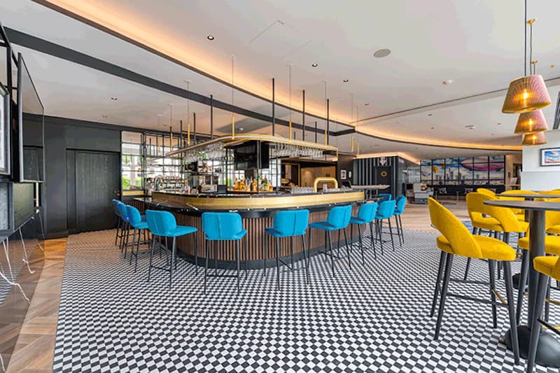 Runde Hotelbar mit blau gepolsterten Hockern, gelben Barstühlen und schwarz-weiß kariertem Boden.
