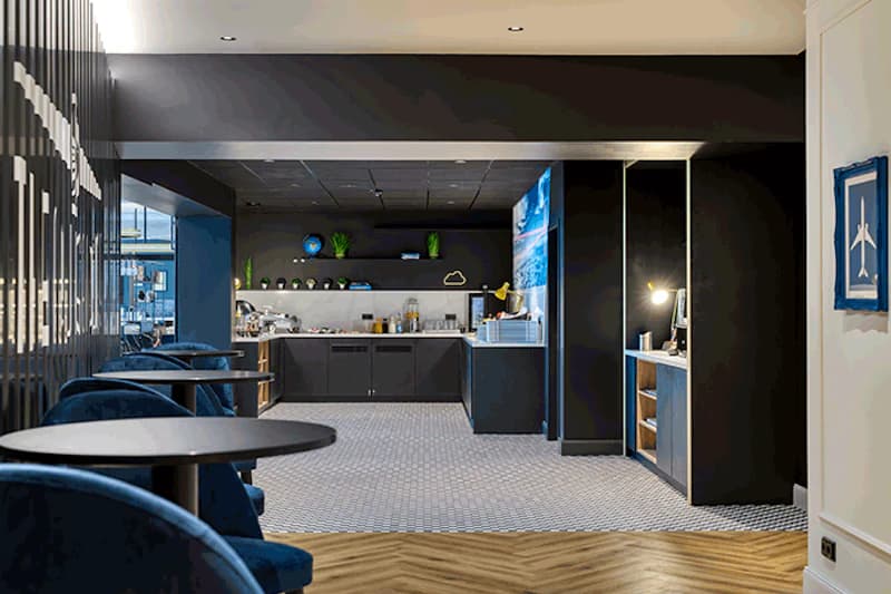 Hotellounge mit runden Tischen, blauen Polstersesseln und Buffetzeile mit Kaffeemaschine und Dekoration.