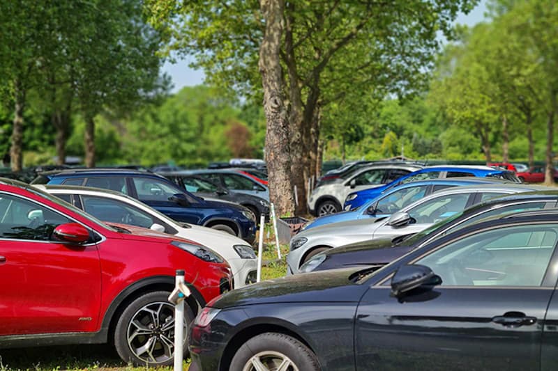 Reihen geparkter Autos auf einem Freiluftparkplatz zwischen Bäumen und Gras.