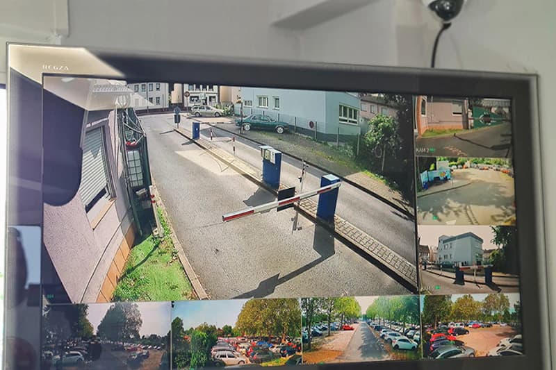 Überwachungsbildschirm mit mehreren Kamerabildern einer Parkplatzeinfahrt mit Schranken und geparkten Autos.