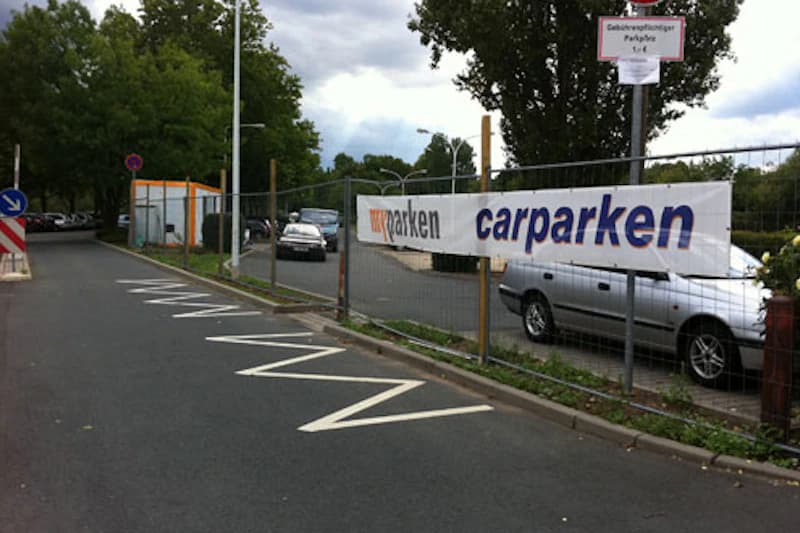 Eingezäunter Parkplatz an Zufahrtsstraße mit Werbebanner carparken, mehreren geparkten Pkw und kleinem Kassenhäuschen.