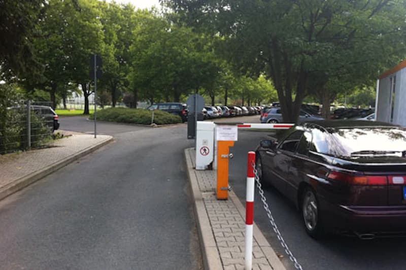 Einfahrt zu Parkfläche mit Schranke, orangener Kassenautomat, dunkles Auto rechts und Reihe geparkter Fahrzeuge hinter Bäumen.