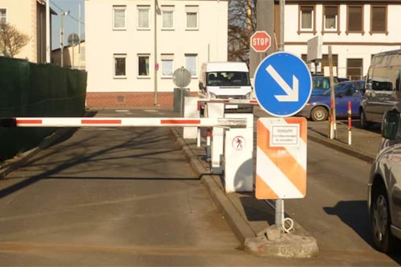 Schranke an Parkplatzeinfahrt mit abgesenkter Barriere, blauer Pfeil rechts unten, Stoppschild und Lieferwagen im Hintergrund.