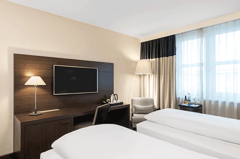 Zweibettzimmer mit zwei Einzelbetten, Schreibtisch mit Fernseher, Tisch- und Stehleuchte, Sessel und Fenster mit Vorhängen.