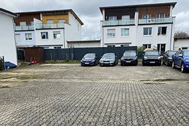 Gepflasterter Parkplatz mit fünf geparkten Autos vor dunklem Zaun und Wohnhäusern bei bewölktem Himmel.