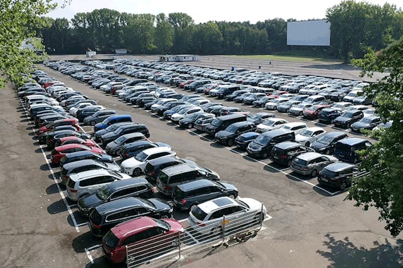 Großer Außenparkplatz mit dicht geparkten Reihen von Autos, Baumreihe im Hintergrund und großer weißer Leinwand.