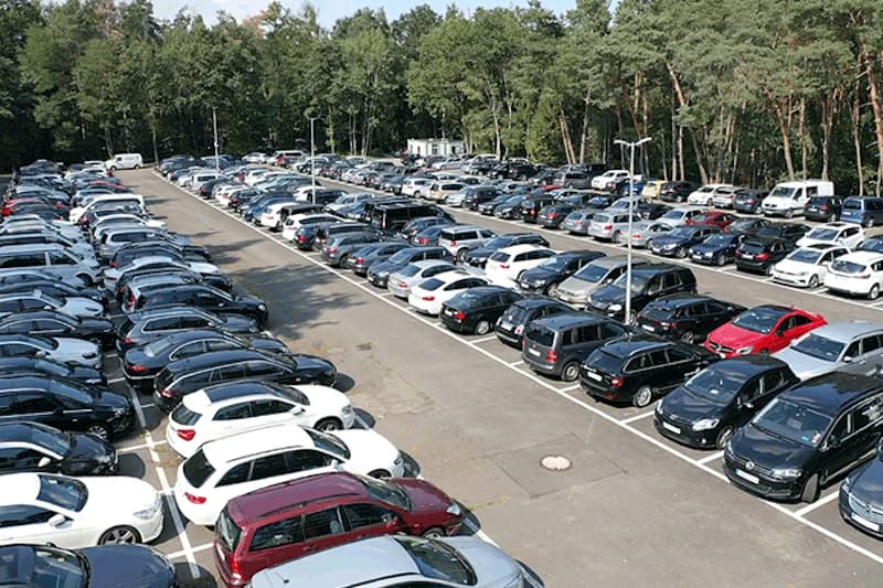 Großer Außenparkplatz mit markierten Stellplätzen, vielen geparkten Fahrzeugen, Beleuchtungspfosten und Baumbestand am Rand.