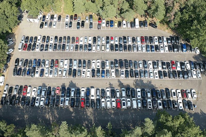 Luftaufnahme eines Parkplatzes mit mehreren dicht geparkten Reihen Autos, Baumgrenze und kleiner Kontrollhütte oben links.