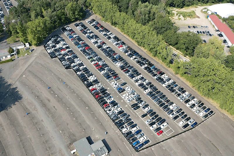 Luftaufnahme eines Freiluftparkplatzes mit mehreren geraden Reihen geparkter Autos neben Baumbestand am Flughafen Frankfurt.