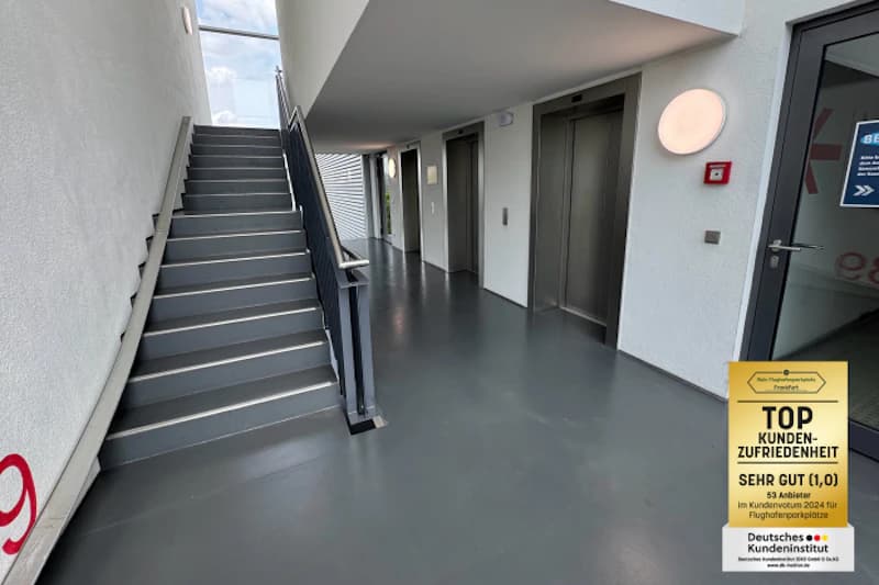 Innenbereich mit Treppe und Handlauf, zwei Aufzügen, Tür, runder Wandlampe und Feueralarm, grauer Boden.