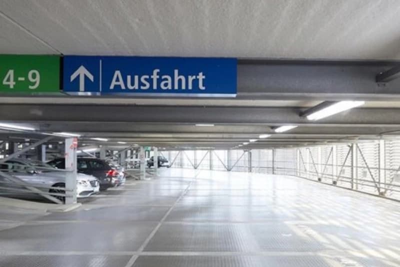 Mehrstöckiges Parkdeck mit blauem Ausfahrt-Schild, geparkten Fahrzeugen links und Metallgeländer rechts.