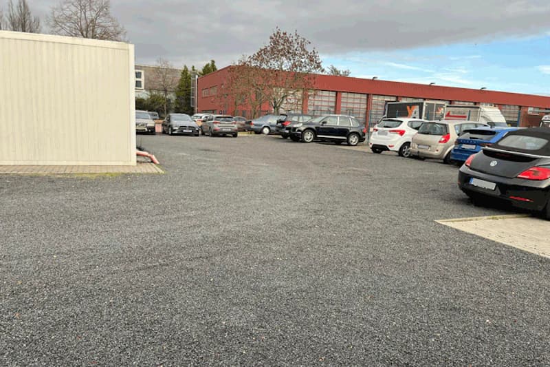 Schotterparkplatz mit geparkten Autos rechts, rotem Industriegebäude im Hintergrund und weißem Container links.