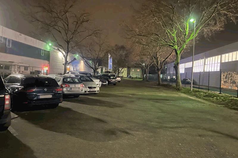 Abendlicher Außenparkplatz mit Reihe geparkter Autos links, Bäumen und beleuchteten Gebäuden rechts.