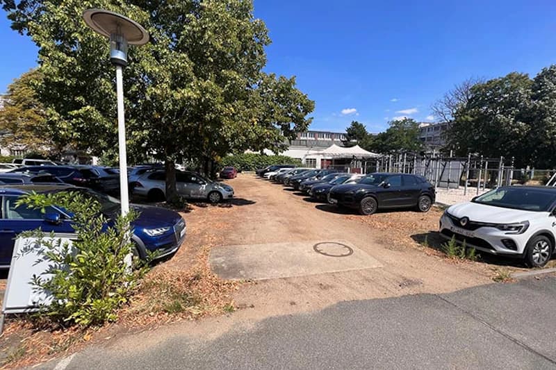Geschotterter Parkplatz mit zwei Reihen geparkter Autos, Baumbestand, Straßenlaterne und Gebäuden im Hintergrund.